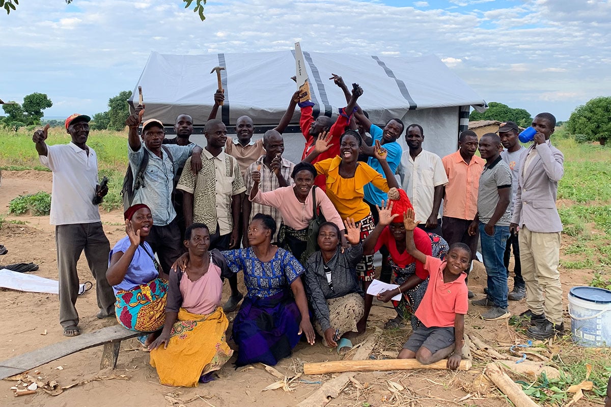 A group of Malawians gather in front of a shelter raising tools in celebration