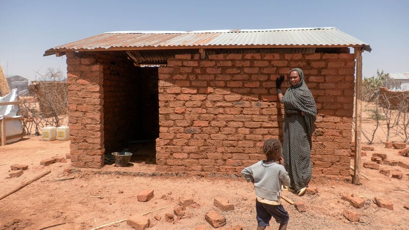 A woman standing outside of a semi durable shelter with a little boy standing next to her, in Chad, after fleeing the Sudan war.