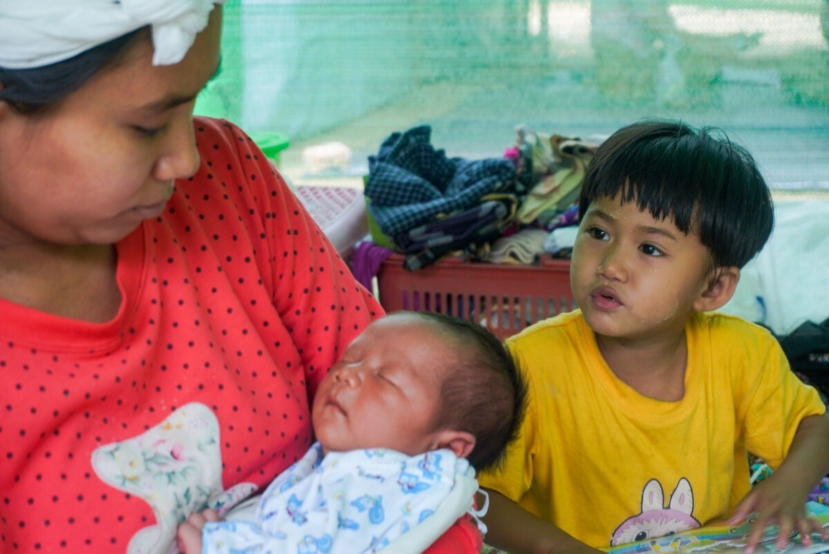 A woman holding her baby with a boy in the background in an emergency shelter.